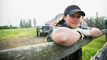 A woman leaning on the rail of a wooden fence with her arms crossed. She is wearing work gloves and baseball cap. A barn and pickup truck are in the background.