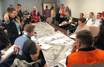 About 15 to 20 people in a room and gathered around a table. They are from the CER, the company, and local authorities working together in unified command to practice a response to an emergency.