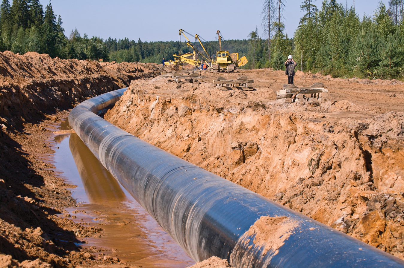 Un pipeline dans une tranchée boueuse, avec des travailleurs et de l’équipement lourd en arrière-plan.