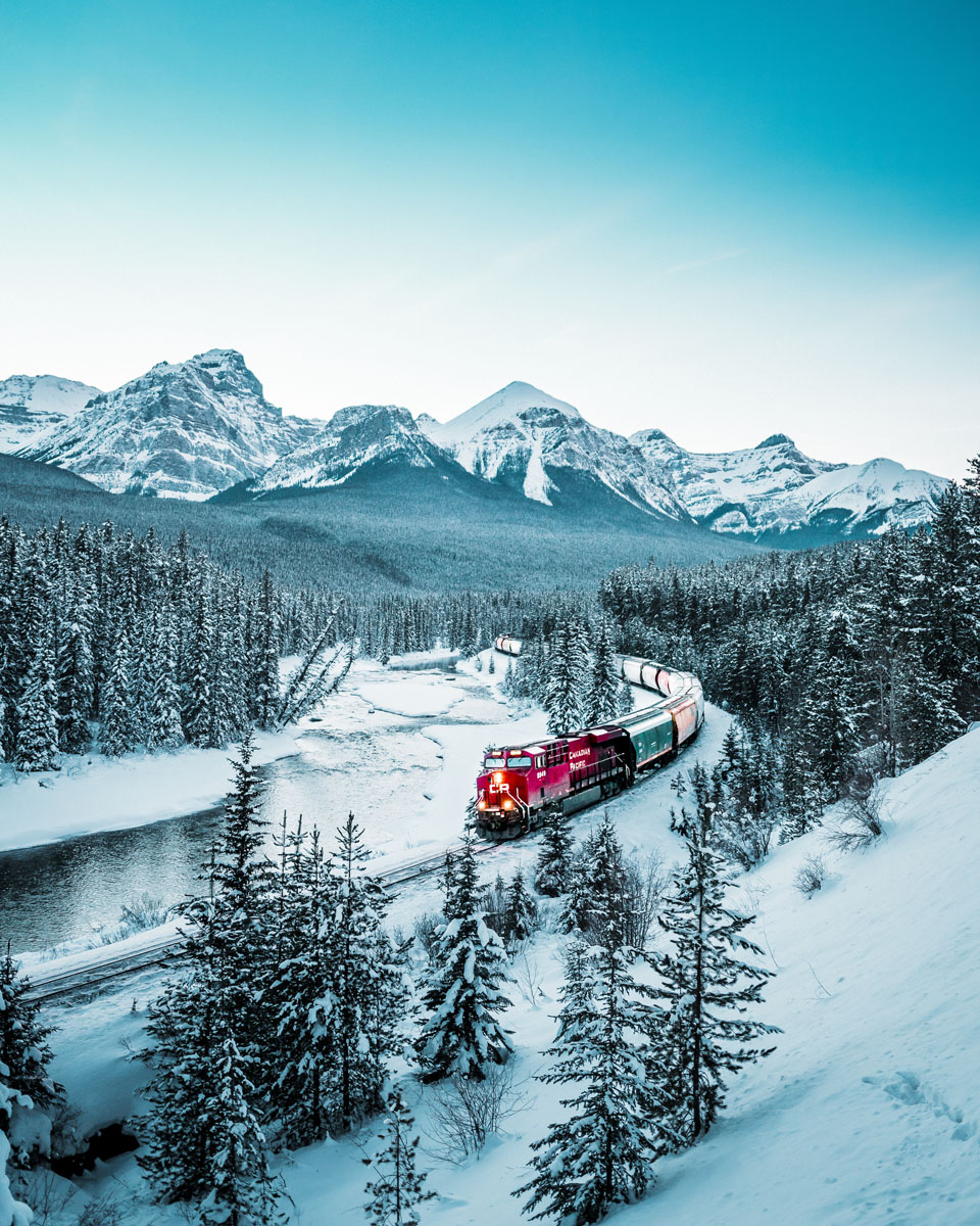 Train moving next to river, in the snow.	Un train qui circule près d’une rivière dans un paysage enneigé.