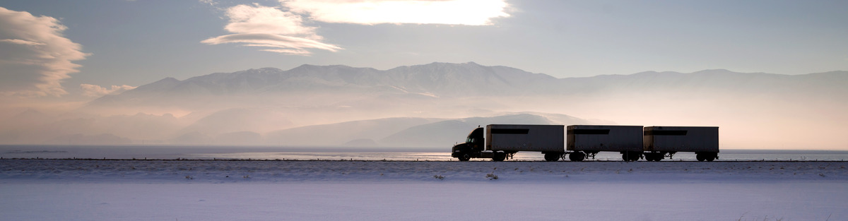 Transport truck drives through snowy prairies with mountains in background.
