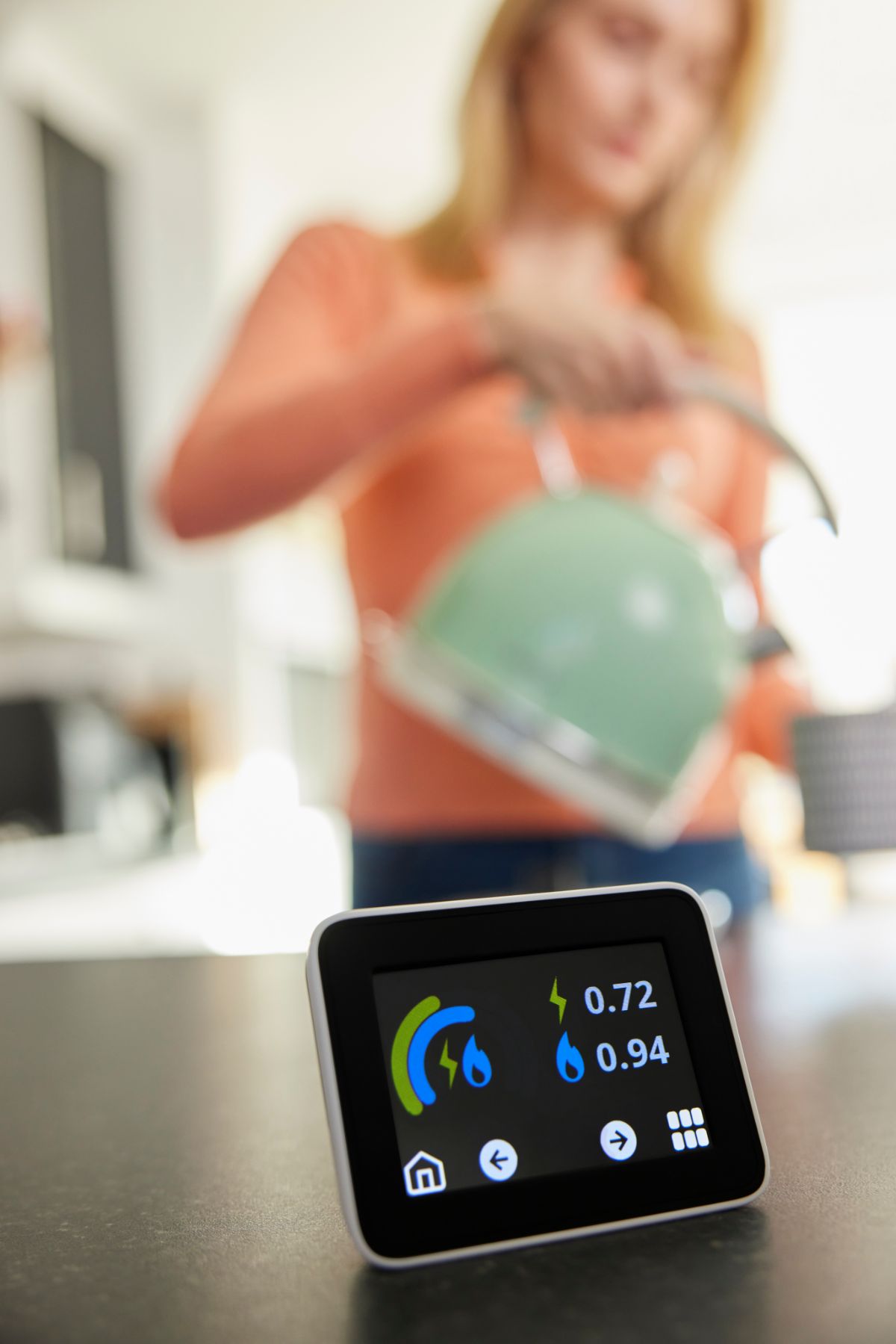 Home energy meter in foreground with woman pouring kettle in background.