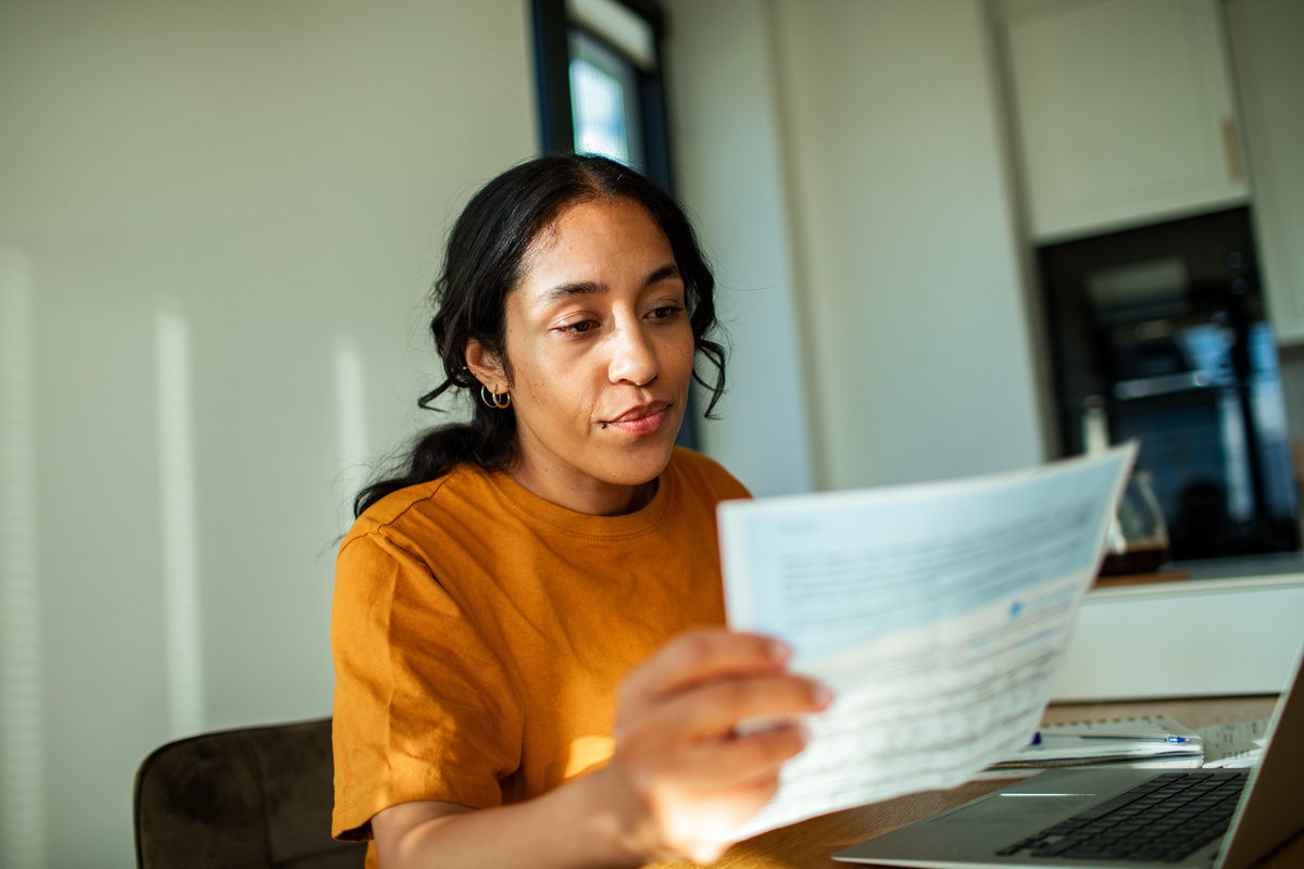 Une jeune femme vêtue d'un t-shirt orange est assise à un bureau devant un ordinateur portable, en train de consulter un document papier, une facture d'électricité.