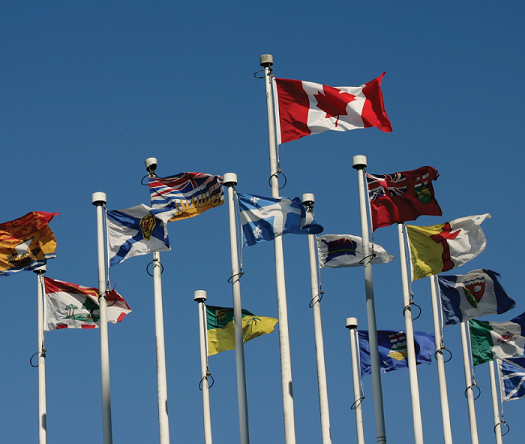 Photo: The Province and Territory flags fly in the wind against a blue sky.
