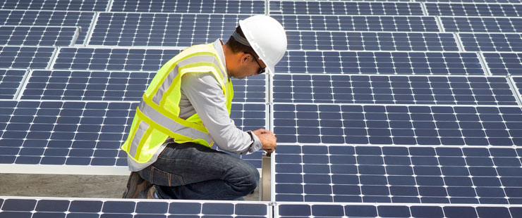 ) A technician in PPE working on solar panels on a building’s roof