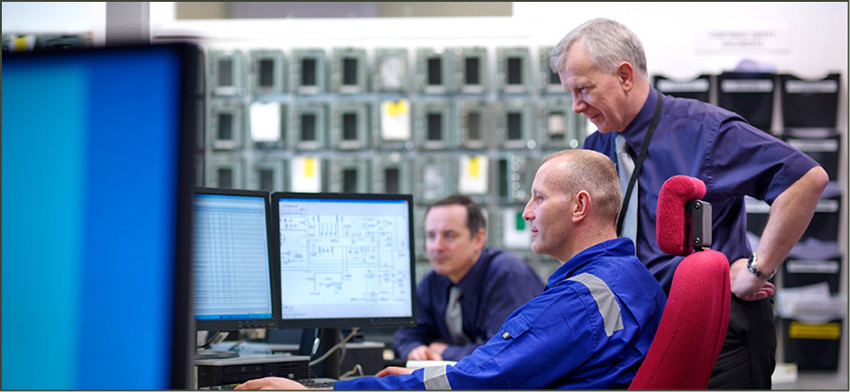Two engineers sit at a desk reviewing documents on a computer screen in a power plant control room.