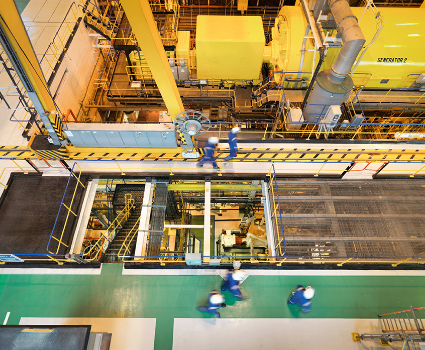 Arial shot of a generator room in a nuclear power plant, engineers walking beside it on a bridge in personal protective equipment.