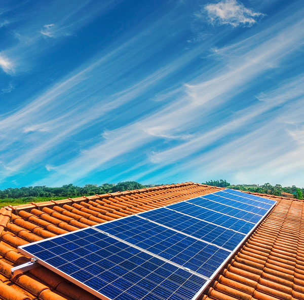 Solar panels on a terracotta tile roof, with green trees and blue sky with wispy clouds in the background.