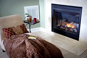 A photo of a chaise lounge with a book and a blanket laid out on it sits in front of a natural gas fireplace.
