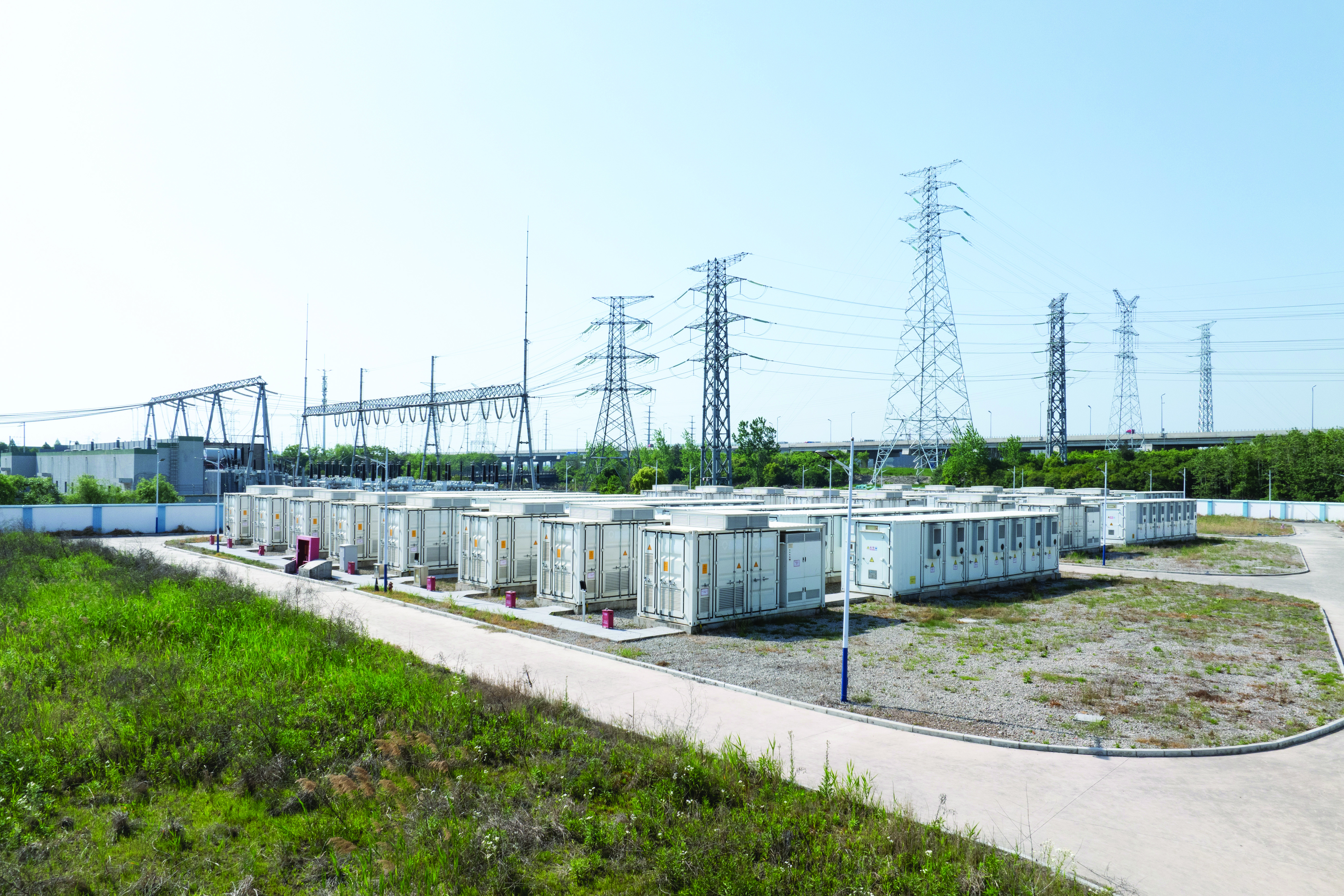 Grey and white energy storage batteries on a gravel lot next to a substation on a partly cloudy day.