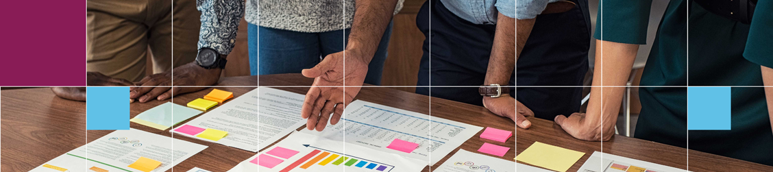 Banner showing various people in an office environment gathered around a desk motioning with their hands at a variety of print out information sheets